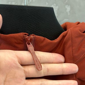 May include: Close-up of a rust-colored jacket zipper with the brand name "ARC'TERYX" visible on the zipper pull. The jacket is hanging on a black hanger.