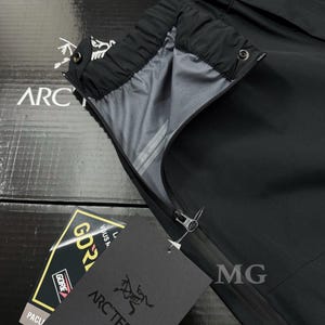 May include: Black outdoor pants with a zipper and a tag that says "ARC'TERYX". The pants have a gray interior lining and a black exterior. The tag also has the word "GORE-TEX" on it. The pants are likely designed for outdoor activities.