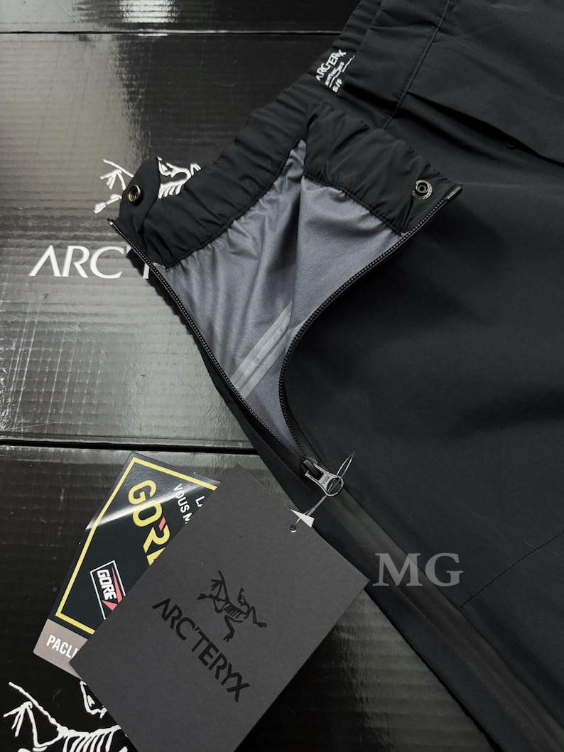 May include: Black outdoor pants with a zipper and a tag that says "ARC'TERYX". The pants have a gray interior lining and a black exterior. The tag also has the word "GORE-TEX" on it. The pants are likely designed for outdoor activities.