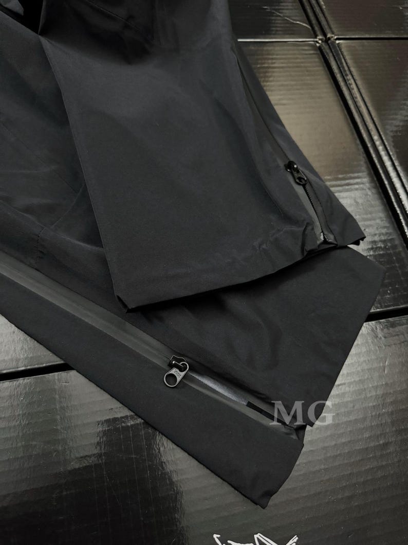 May include: Black waterproof pants with zippers along the sides. The pants are made of a smooth, matte material. The letters "MG" are visible on the pants.