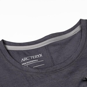 May include: Close-up of a dark gray crew neck t-shirt. The shirt features a gray neckline and the brand name "ARC'TERYX" printed on the chest. The shirt has a drawstring detail and is made of a lightweight material.