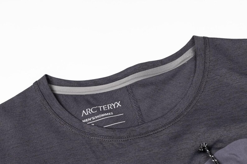 May include: Close-up of a dark gray crew neck t-shirt. The shirt features a gray neckline and the brand name "ARC'TERYX" printed on the chest. The shirt has a drawstring detail and is made of a lightweight material.