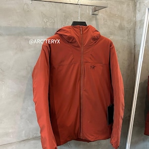 May include: A rust-colored hooded jacket with a full-length zipper and a small logo on the chest. The jacket is hanging on a metal rack against a textured gray wall. The brand name "ARC'TERYX" is visible in the image.