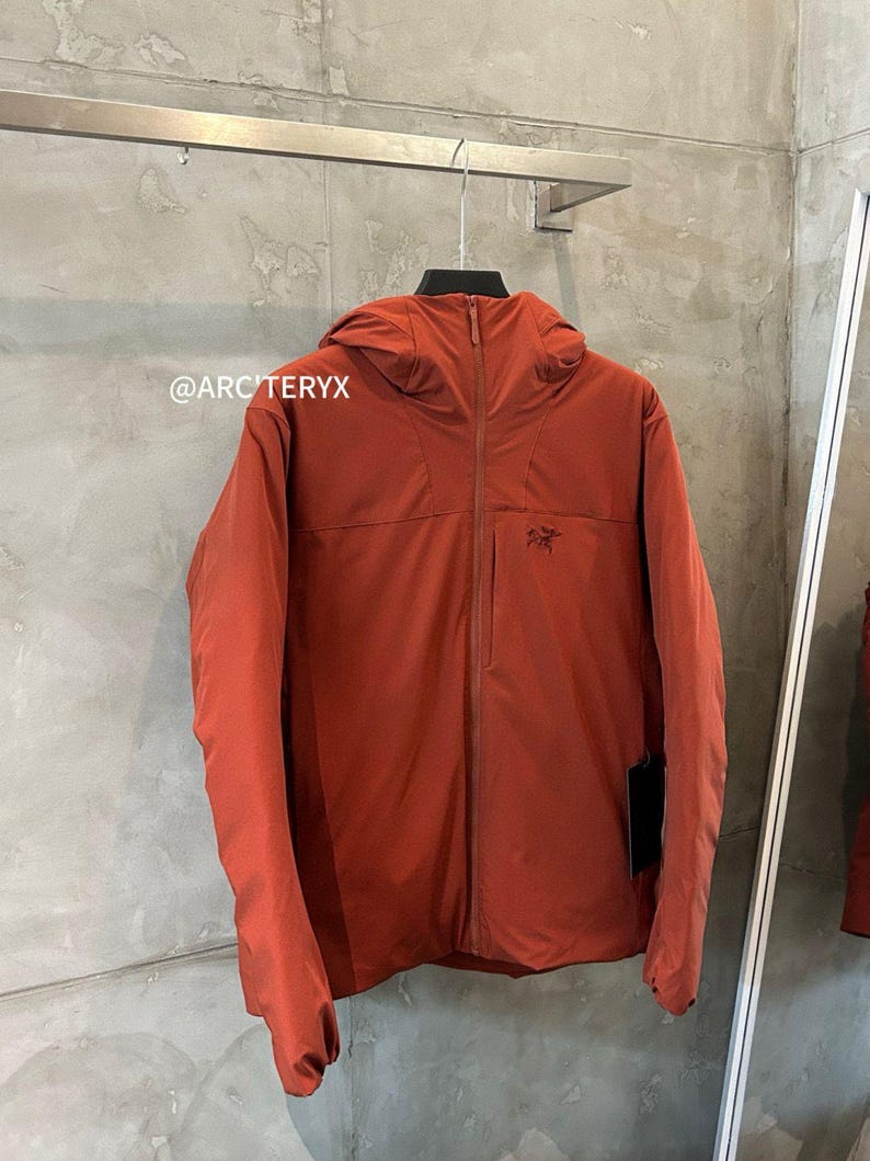 May include: A rust-colored hooded jacket with a full-length zipper and a small logo on the chest. The jacket is hanging on a metal rack against a textured gray wall. The brand name "ARC'TERYX" is visible in the image.