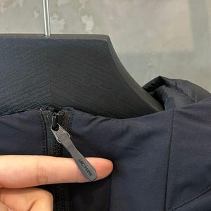 May include: Close-up of a navy blue jacket with a black zipper. The zipper pull has the brand name "ARC'TERYX" printed on it. The jacket is hanging on a black hanger.