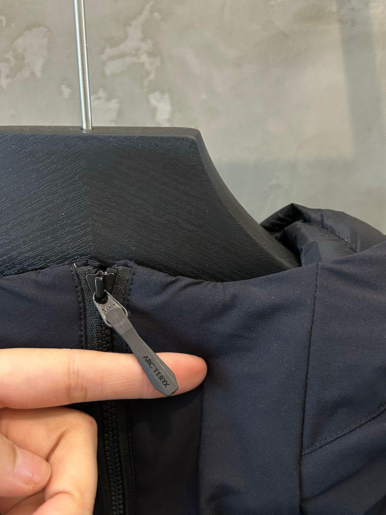 May include: Close-up of a navy blue jacket with a black zipper. The zipper pull has the brand name "ARC'TERYX" printed on it. The jacket is hanging on a black hanger.