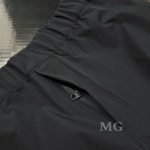 May include: Close-up of black athletic pants with a zippered pocket. The pants have an elastic waistband and a smooth, matte finish. The letters "MG" are printed on the pants.