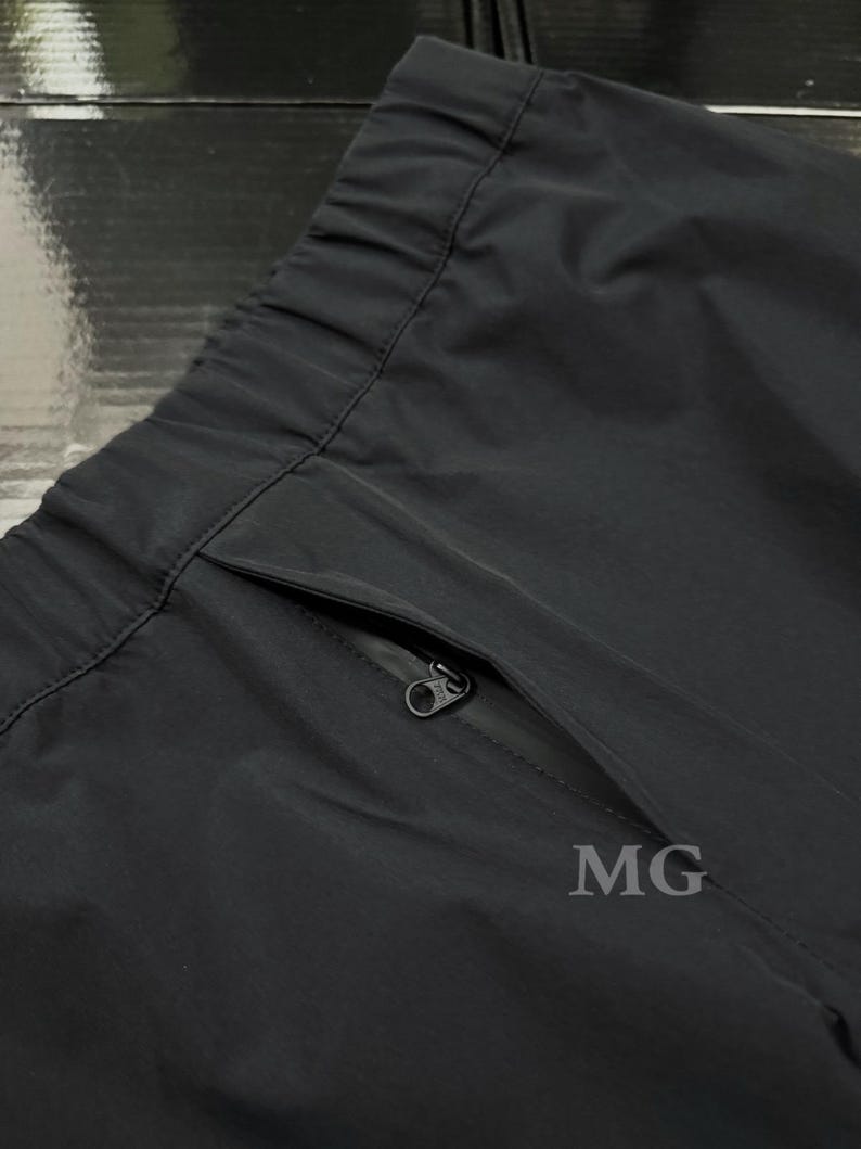 May include: Close-up of black athletic pants with a zippered pocket. The pants have an elastic waistband and a smooth, matte finish. The letters "MG" are printed on the pants.