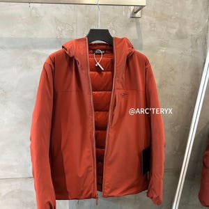 May include: A rust-colored hooded jacket with a zipper closure, hanging on a black hanger. The jacket has a quilted interior and the text "@ARC'TERYX" is visible on the right side.