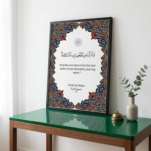 May include: A framed wall art print featuring a white center with a black floral icon, Arabic calligraphy, and the English text: "And We sent down from the rain-laden clouds abundant, pouring water." Surah An-Naba 14. The border has blue and red patterns.