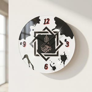 Op de afbeelding: Een ronde wandklok met een witte achtergrond en zwarte inktachtige spetters. Het midden toont een zwart vierkant met een in elkaar grijpend geometrisch sterpatroon en Arabische kalligrafie. De cijfers 3, 6, 9 en 12 zijn in het rood.