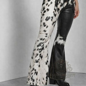 Op de afbeelding: Uitlopende leren broek, één been met een koeienhuidpatroon van zwarte vlekken op wit, de andere van effen zwart leer. De broek heeft een hoge taille en een uitlopende pijp. De merknaam "LEATHERSOCIETY" is zichtbaar.