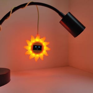 May include: A bright orange sun-shaped light fixture with a central digital display showing a simple face with two eyes and a smile. The sun light is suspended from a flexible black gooseneck lamp arm that features a cylindrical black light head.