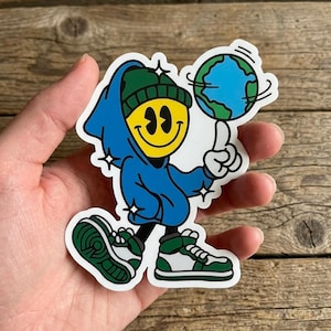 May include: A sticker featuring a cartoon character with a yellow smiley face wearing a blue hoodie and green beanie. The character is balancing a globe on one finger and wearing green and white sneakers. The sticker has a white border and star accents.