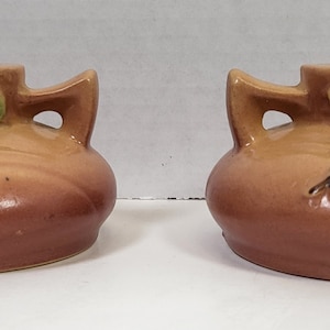 May include: Two ceramic candle holders with a brown and tan gradient. Each has a small green leaf and a tan stem design. The candle holders have two handles and a circular opening for a candle.
