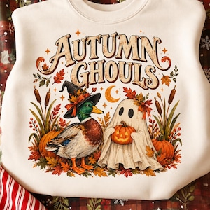 Peut inclure: Un sweat-shirt à col rond couleur crème portant l'inscription "AUTUMN GHOULS" dans une police stylisée. Le motif représente un canard colvert portant un chapeau de sorcière et un fantôme tenant une citrouille, entourés de massettes et de feuilles d'automne.