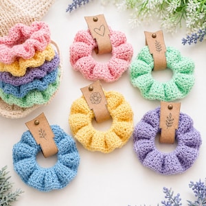 May include: A collection of colorful, handmade crochet scrunchies in various pastel shades, including pink, yellow, green, blue, and purple. Each scrunchie has a leather tag with a decorative design. Some are stacked, others are displayed individually.