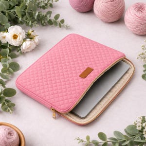 Può includere: Una custodia trapuntata rosa per computer portatile con motivo a rombi e una piccola toppa rettangolare marrone sul davanti. La custodia è parzialmente aperta, rivelando un computer portatile argentato e una cerniera metallica dorata.