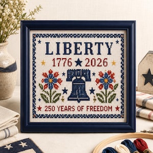 May include: A framed cross-stitch sampler featuring the word LIBERTY in blue, the years 1776 and 2026, a central blue Liberty Bell, two floral motifs in red, blue, and green, and the text 250 YEARS OF FREEDOM, all enclosed in a dark blue frame.