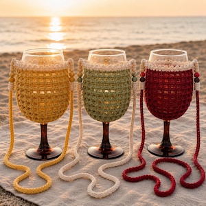 May include: Three wine glasses with crochet covers in yellow, sage green, and deep red. Each cover features a grid pattern, a cream-coloured top border, and a matching crochet strap adorned with wooden beads on the sides for carrying.