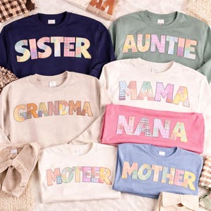 May include: A selection of crew neck sweatshirts and t-shirts in various colours, each displaying a family-related word in a patchwork design. The words include "SISTER", "AUNTIE", "GRANDMA", "MAMA", "NANA", "MOTHER", and "MOTHER".