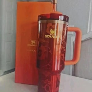 May include: A red and gold Stanley travel mug with a pineapple-shaped topper and an orange handle. The mug has a floral design and the Stanley logo. An orange box is behind the mug.