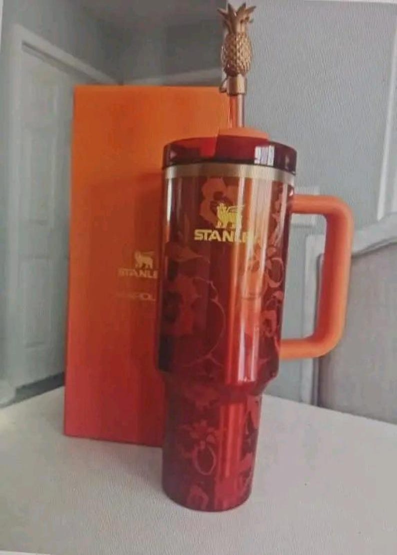 May include: A red and gold Stanley travel mug with a pineapple-shaped topper and an orange handle. The mug has a floral design and the Stanley logo. An orange box is behind the mug.