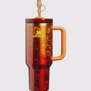 May include: A red and orange Stanley tumbler with a handle and a pineapple-shaped topper. The tumbler features a floral design and the Stanley logo in gold. The handle and topper are orange.