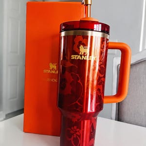 May include: A red and orange Stanley tumbler with a floral design and a gold pineapple topper. The tumbler has a handle and the Stanley logo. An orange box is behind the tumbler.