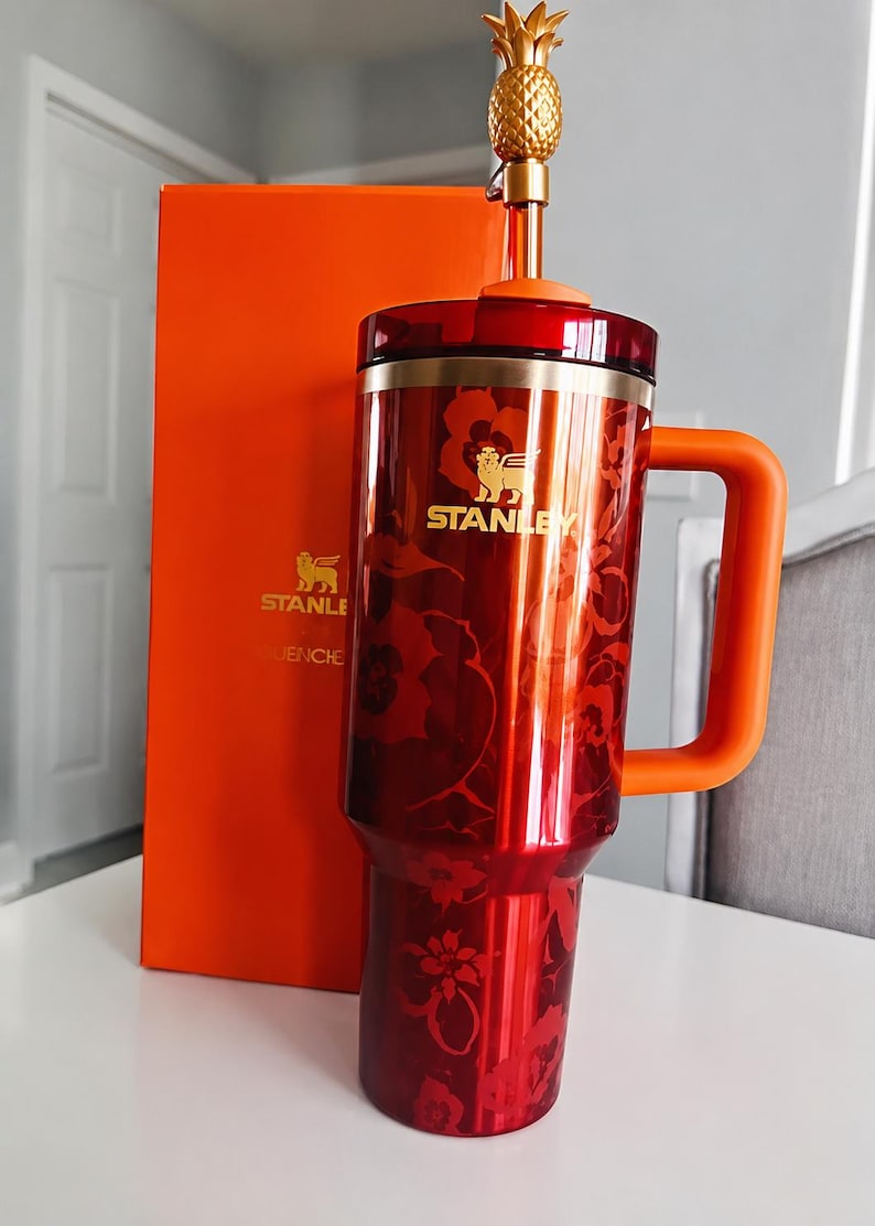 May include: A red and orange Stanley tumbler with a floral design and a gold pineapple topper. The tumbler has a handle and the Stanley logo. An orange box is behind the tumbler.