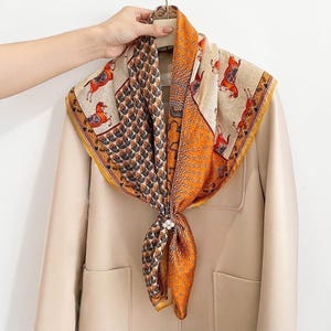 May include: A square scarf featuring a multi-panel design with orange, beige, and brown tones. The pattern includes illustrations of horses, geometric shapes, and dotted textures. The scarf is secured with a small, circular, white floral-shaped clip.