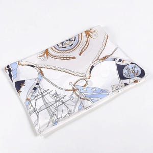 May include: A folded square scarf featuring a nautical print on a white background. The design includes illustrations of sailing ships, ropes, compasses, and flags in shades of navy blue, light blue, and tan, with anchor motifs on the dark blue flags.