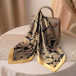 Könnte beinhalten: Ein schwarzer und goldener Seidenschal mit Paisley-Muster, der über einer grauen Handtasche drapiert ist. Der Schal hat einen goldenen Rand und ein detailliertes Design aus wirbelnden Paisley-Formen. Die Handtasche hat einen oberen Griff und eine rechteckige Form.