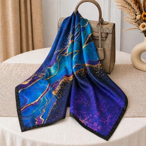 May include: A square silk scarf featuring a vibrant blue and purple abstract marble pattern with gold-colored vein accents and a black border, draped over a structured metallic gold handbag with a top handle and a rectangular frame.