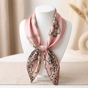 May include: A square silk scarf in a soft pink hue featuring a decorative pattern of gold-toned chains, brown leopard print sections, and intricate dark brown floral motifs. The scarf is tied in a knot at the center, displaying a smooth finish.