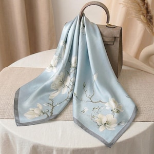 May include: A light blue square scarf featuring a white floral pattern of magnolia blossoms on thin brown branches, finished with a dark gray border, draped over a structured taupe handbag with a top handle and a metallic clasp closure.