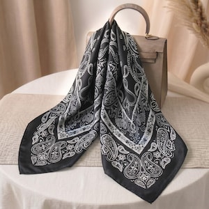 May include: A square black scarf featuring a white paisley pattern with intricate floral and teardrop motifs. The scarf is draped over a structured beige handbag with a single top handle, showcasing a smooth fabric texture and a classic border design.