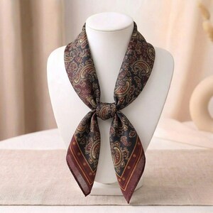 May include: A square scarf featuring a dark brown and black paisley pattern with intricate gold and cream details. The scarf is tied in a knot at the centre, displaying a wide border with thin stripes and small star motifs along the bottom edges.