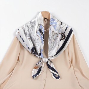 May include: A square silk scarf featuring a nautical theme with illustrations of ships, anchors, and ropes in shades of navy blue, light blue, and white. The scarf is tied in a knot and draped over a plain beige long-sleeved button-down shirt.