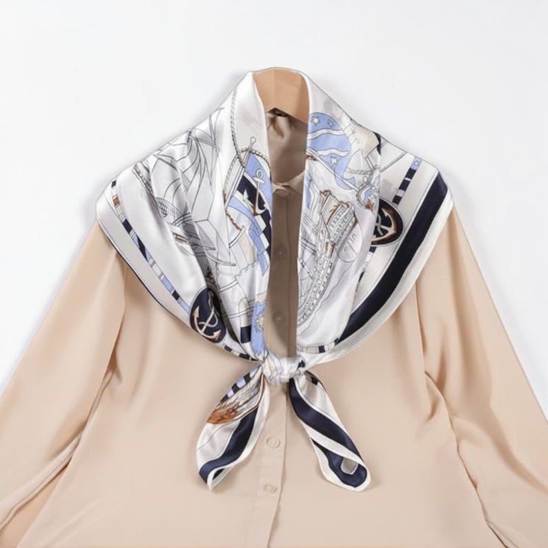 May include: A square silk scarf featuring a nautical theme with illustrations of ships, anchors, and ropes in shades of navy blue, light blue, and white. The scarf is tied in a knot and draped over a plain beige long-sleeved button-down shirt.