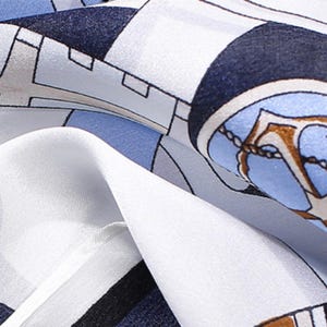 May include: A smooth fabric scarf featuring a geometric pattern in shades of navy blue, light blue, white, and grey. The design includes circular motifs with gold-toned anchor illustrations and chain details printed across the surface.