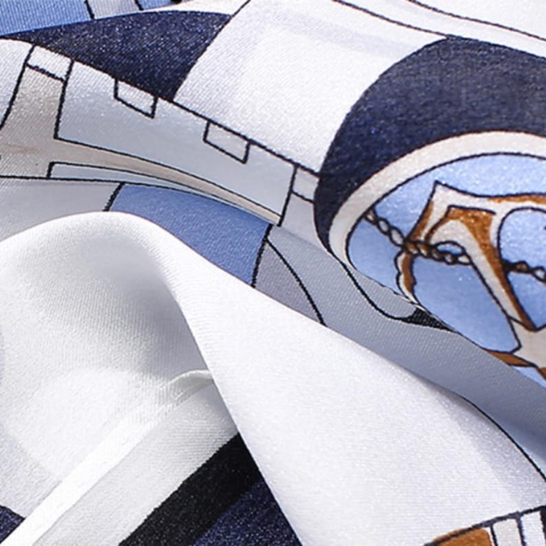 May include: A smooth fabric scarf featuring a geometric pattern in shades of navy blue, light blue, white, and grey. The design includes circular motifs with gold-toned anchor illustrations and chain details printed across the surface.