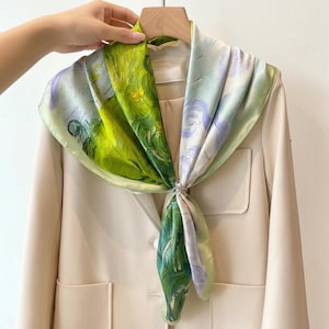 May include: A square silk scarf featuring an impressionist-style print with vibrant shades of lime green, forest green, and soft lavender, tied with a small silver-toned metal ring at the center, draped over a beige blazer on a wooden hanger.