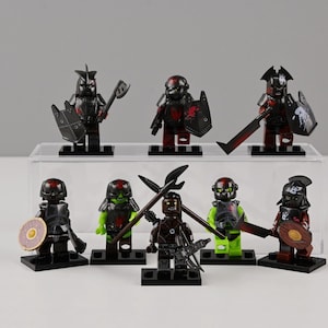 Puede incluir: Una colección de ocho figuras de guerreros en miniatura, cada una con armadura y armas únicas. Las figuras son principalmente negras con detalles rojos, algunas con escudos y hachas. Se exhiben sobre bases negras individuales.