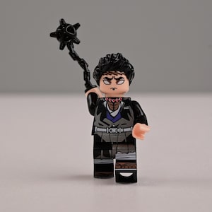 以下が含まれることがあります： 黒い髪と険しい表情をしたキャラクターのレゴフィギュア。黒いトゲのある武器を持っています。フィギュアは、白いベルトと茶色のアクセントが付いた黒とグレーの衣装を着ています。背景は無地のグレーです。
