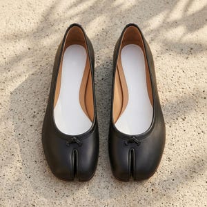 Könnte beinhalten: Ein Paar schwarze Ballerinas aus Leder mit geteilter Zehenpartie, glatter Oberfläche, einem kleinen dekorativen Knoten auf dem Vorderblatt und einer weißen Innensohle. Die Schuhe haben ein markantes Design mit Schlitz für den großen Zeh.