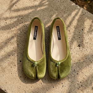 May include: A pair of olive green velvet split-toe flats featuring a small decorative knot detail at the front. The interior insoles are cream-colored and clearly display the brand name OKLEIBO printed in black text on the heel area.