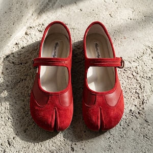 Op de afbeelding: Een paar rode Mary Jane-schoenen met een split-teenontwerp. De schoenen hebben een band over de bovenkant met een gespsluiting. De schoenen zijn gemaakt van suède en leer. De binnenkant van de schoen heeft de tekst "River July".