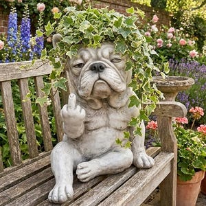Puede incluir: Estatua de bulldog gris con una planta verde en la cabeza, sentada en un banco de madera. La estatua está mostrando el dedo medio. El fondo presenta un jardín con flores y un muro de ladrillos.