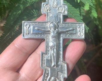 May include: A silver-colored Orthodox cross pendant with a detailed depiction of the crucifixion. The cross features intricate engravings and a small figure of Jesus Christ. The pendant is held in a hand against a backdrop of green foliage.
